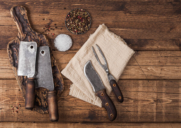Vintage hatchets for meat on wooden chopping board with salt and pepper on wooden table background with linen towel and fork and knife. Top view