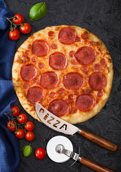 Fresh round baked Pepperoni italian pizza with wheel cutter and knife with tomatoes and basil on black background. Top view