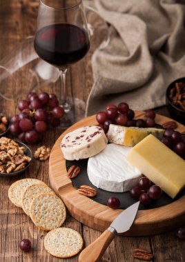 Tahtasında çeşitli peynirler ve ahşap masa zemininde üzümler olan bir bardak kırmızı şarap. Mavi Stilton, Kırmızı Leicester ve peynirli peynir ve keten mutfak bezli bıçak..