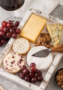 Bir bardak kırmızı şarap, ahşap kutuda çeşitli peynirler ve hafif arka planda üzümler. Mavi Stilton, Kırmızı Leicester ve Cheddar ve bıçaklı Brie Cheese..