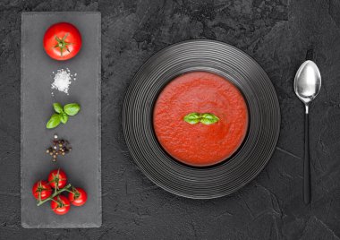 Siyah bir tabak kremalı domates çorbası ve siyah zemin üzerinde kaşık, taş doğrama tahtası, çiğ domates, biber ve tuz.. 