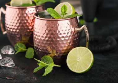 Bakır bardakta Moskova katırı kokteyli, kireç, nane ve ahşap sıkacağı, mutfak arka planında da buz küpleri..