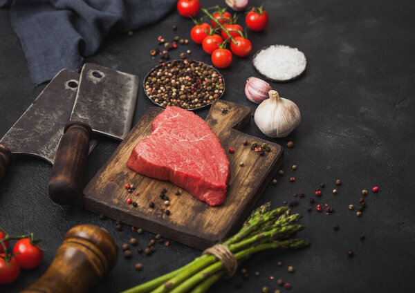 Slice of Raw Beef sirlion steak on wooden chopping board with tomatoes,garlic and asparagus tips and meat hatchet on dark kitchen table background.