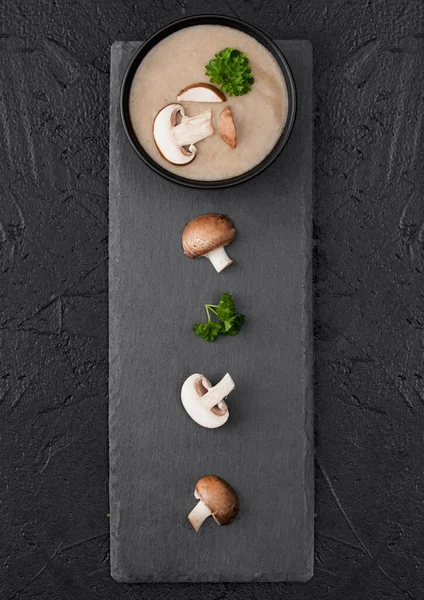 用黑石板和新鲜蘑菇在黑色背景上做的奶油栗子香菇汤的黑碗盘 — 图库照片