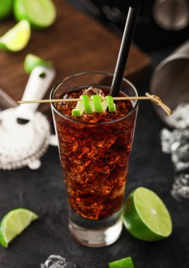 Buzlu viski bardağında Küba Libre kokteyli bambu çubuğunda limon kabuğu, saman ve siyah masa arkasında taze limon. Titretici ve süzgeç büfede. Üst görünüm
