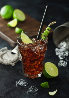 Küba Libre kokteyli ağır kristal bardakta buz ve limon dilimi ve kabuğu ile, saman ve taze limon ile siyah masa arkasında süzgeç ile.