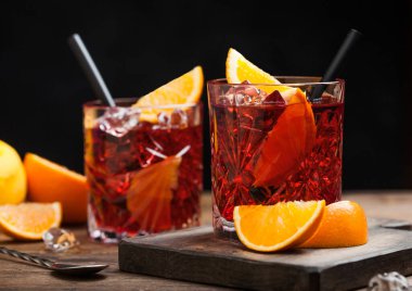 Negroni cocktail in crystal glasses with orange slice and fresh raw oranges on chopping board with strainer on wood background. Macro clipart