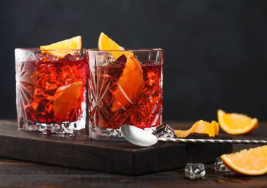 Kristal bardaklarda Negroni kokteyli, portakal dilimi ve tahta üzerinde taze çiğ portakallar, ahşap arka planda kaşık..