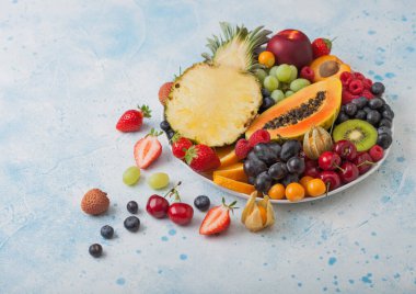Taze çiğ organik yaz meyveleri ve mavi mutfak arka planında beyaz tabakta egzotik meyveler. Ananas, papaya, üzüm, nektarin, portakal, kayısı, kivi, armut, lychees, kiraz ve physalis. Üst görünüm