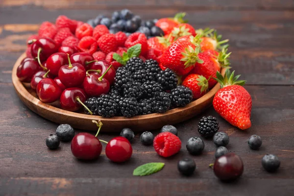 Fresh organic summer berries mix in round wooden tray on dark wooden table background. Raspberries, strawberries, blueberries, blackberries and cherries. Top view