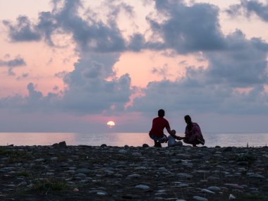 Mutlu aile - baba, anne, bebek evlat günbatımı deniz üzerinde siyah kum plaj sörf bakın. Çocuklu etkin ebeveynler ve insanlar açık hava etkinliği tropikal yaz tatillerini.