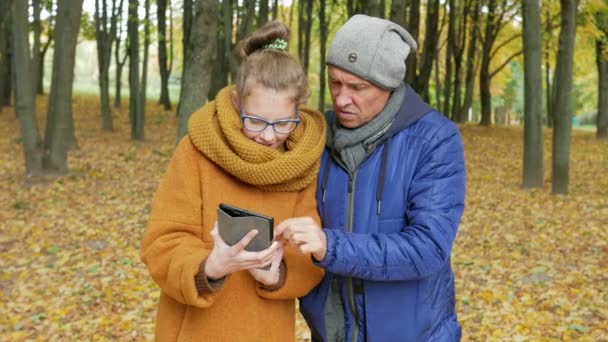 Daughter teenager teaches his father to work with the tablet in the autumn park. Dad listens carefully and repeats the daughter of her touch display — Stock Video