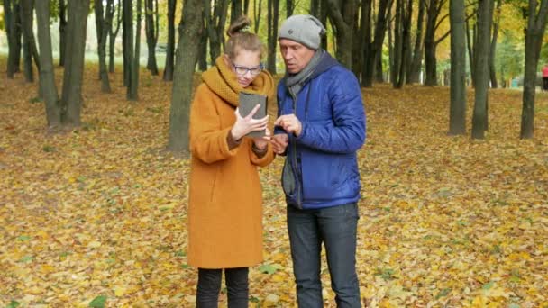 Tiener dochter leert zijn vader om te werken met de tablet in het najaar park. Vader luistert aandachtig en herhaalt de dochter van haar touch-display — Stockvideo