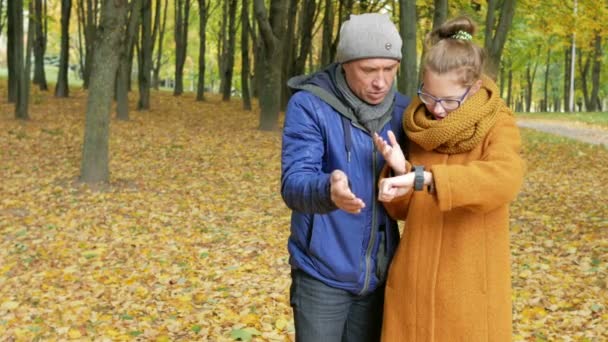 Hija adolescente enseña a su padre a trabajar con el reloj inteligente en el parque de otoño. Papá escucha atentamente y repite a la hija de su pantalla táctil — Vídeos de Stock