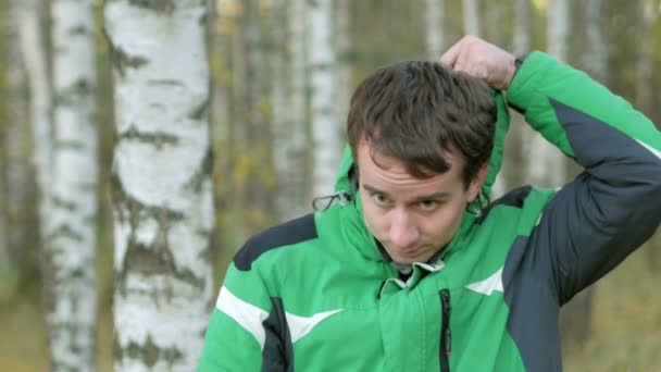 Jeune homme beau porte une cagoule dans le parc froid d'automne. Il regarde dans la caméra. Équiper une veste de sport verte chaude — Video