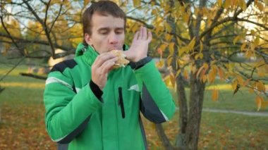 Gün batımında tavuk burger yemek sonbahar ormandaki bir genç adam. Sıcak ceket ve güzel yeşil ağaçlar içinde belgili tanımlık geçmiş