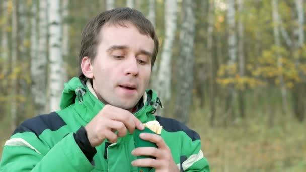 Un joven en el bosque de otoño comiendo papas fritas. Chaqueta cálida y hermosos árboles verdes en el fondo — Vídeos de Stock