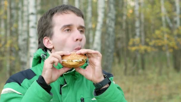En ung man i skogen hösten äter en hamburgare med kyckling. Varm jacka och vackra gröna träden i bakgrunden — Stockvideo