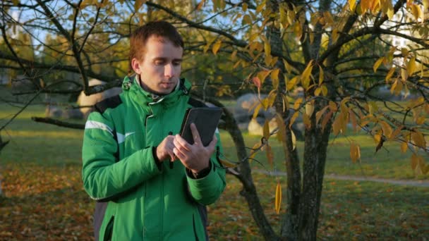Un hombre comprueba los mensajes en las redes sociales en la tableta PC en el parque de otoño al atardecer. Chaqueta deportiva verde y trabajo atento del dispositivo — Vídeos de Stock