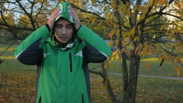 Young handsome man wears a hood in cold autumn park. He looks into the camera. Equip a green sports jacket warm — Stock Video