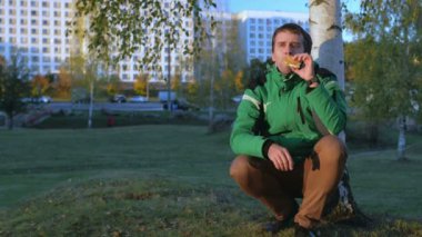 Gün batımında tavuk burger yemek sonbahar ormandaki bir genç adam. Sıcak ceket ve güzel yeşil ağaçlar içinde belgili tanımlık geçmiş