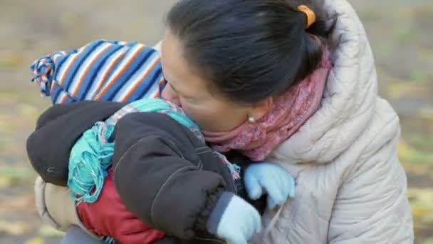 Hermoso bebé está jugando en el parque de otoño con su madre sobre las hojas caídas. El niño está calurosamente vestido con un traje y un sombrero con una bufanda, el niño alrededor de un año — Vídeos de Stock