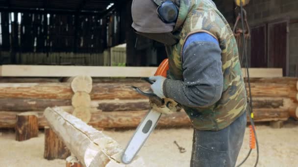 Man maakt krullend snijden hout kettingzaag. Het logboek zal deel uitmaken van de toekomst van het houten huis. Beschermend gezichtsmasker op het gezicht van de bouwer en een heleboel zaagsel. — Stockvideo
