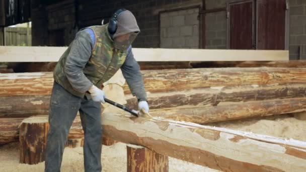 Trabajador de la construcción corta una ranura en el árbol para un futuro hogar. Máscara protectora y auriculares en la cabeza del constructor. Hangar con una parte de la futura casa en el fondo — Vídeos de Stock