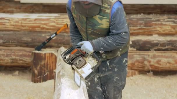 Man maakt krullend snijden hout kettingzaag. Het logboek zal deel uitmaken van de toekomst van het houten huis. Beschermend gezichtsmasker op het gezicht van de bouwer en een heleboel zaagsel. — Stockvideo