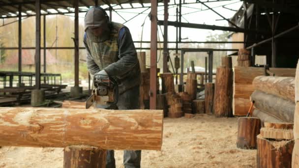 L'homme coupe une tronçonneuse pour sa future maison. Masque facial protecteur sur le visage du constructeur et beaucoup de sciure de bois. Hangar avec une partie de la future maison sur le fond — Video