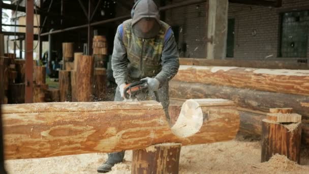 Man skär trä motorsåg för framtida hem. Skyddande ansiktsmask på framsidan byggaren och en hel del sågspån. Hangar med en del av den framtida hem i bakgrunden — Stockvideo
