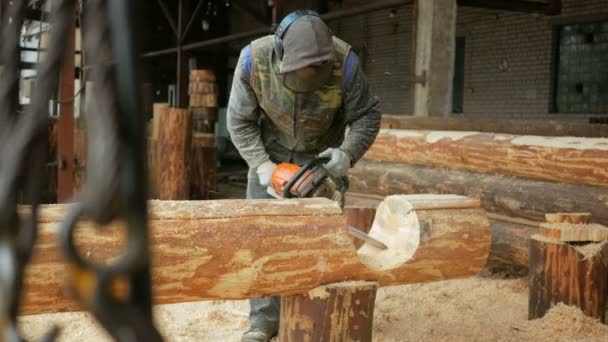 L'uomo taglia la motosega di legno per la futura casa. Maschera protettiva sul viso del costruttore e un sacco di segatura. Hangar con una parte della futura casa sullo sfondo — Video Stock