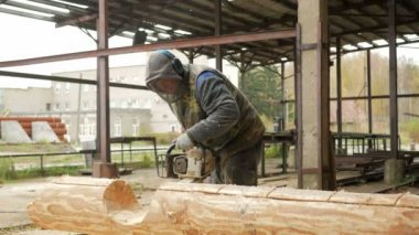 Adam ahşap testere gelecekteki ev için keser. Builder ve bir sürü talaş yüzünde koruyucu yüz maskesi. Hangar gelecekteki ev arka planda bir bölümüyle