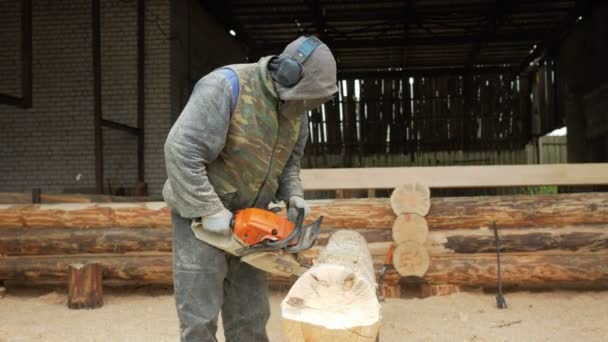 Man snijdt hout kettingzaag voor toekomstige huis. Beschermend gezichtsmasker op het gezicht van de bouwer and alot of zaagsel. Loods met een deel van de toekomstige huis op de achtergrond — Stockvideo
