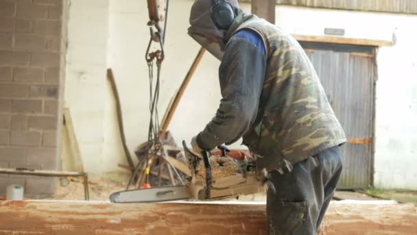 Человек режет деревянную бензопилу для будущего дома. Защитная маска на лице строителя и много опилок. Ангар с частью будущего дома на заднем плане — стоковое видео