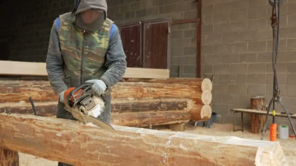 Чоловік вирізає дерев'яну бензопилу для майбутнього будинку. Захисна маска для обличчя будівельника і багато тирси. Ангар з частиною майбутнього будинку на фоні — стокове відео