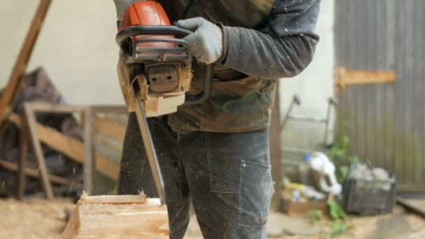 Homem corta motosserra de madeira para casa futura. Máscara protetora no rosto do construtor e muita serragem. Hangar com uma parte da futura casa em segundo plano — Vídeo de Stock