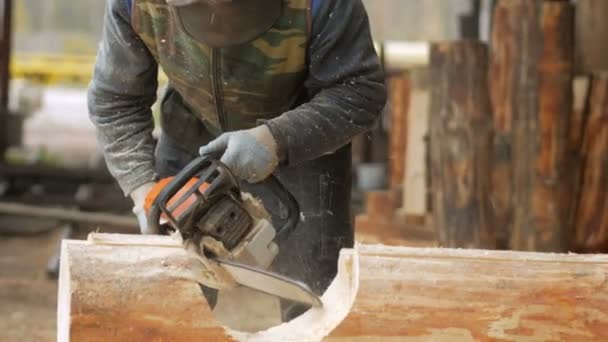 Homem corta motosserra de madeira para casa futura. Máscara protetora no rosto do construtor e muita serragem. Hangar com uma parte da futura casa em segundo plano — Vídeo de Stock