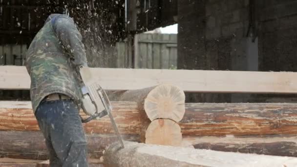 Человек режет деревянную бензопилу для будущего дома. Защитная маска на лице строителя и много опилок. Ангар с частью будущего дома на заднем плане — стоковое видео