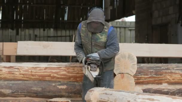 Ο άνθρωπος κόβει ξύλο αλυσοπρίονο για μελλοντικό σπίτι. Μάσκα προστασίας προσώπου εκ πρώτης όψεως ο οικοδόμος και πολλή πριονίδι. Υπόστεγο με ένα μέρος του μέλλοντος στο σπίτι στο παρασκήνιο — Αρχείο Βίντεο