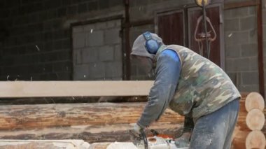 İnşaat işçisi için gelecekteki ev ahşap testere keser. Koruyucu maske ve kulaklık builder ve bir sürü talaş kafasına. Hangar gelecekteki ev arka planda bir bölümüyle