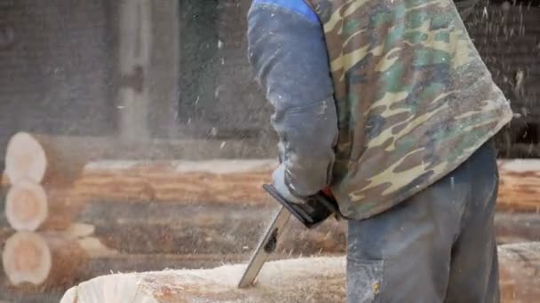 Man cuts wood chainsaw for future home. Protective face mask on the face of the builder and a lot of sawdust. Hangar with a part of the future home on the background — Stock Video