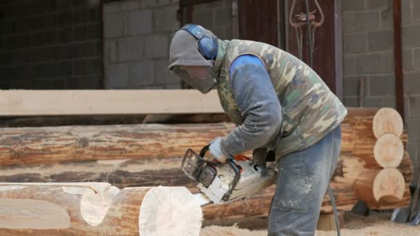 Construction worker cuts wood chainsaw for future home. Protective mask and headphones on the head of the builder and a lot of sawdust. Hangar with a part of the future home on the background — Stock Video