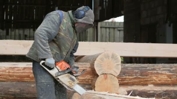 Εργαζόμενος κατασκευή κόβει ξύλο αλυσοπρίονο για μελλοντικό σπίτι. Προστατευτική μάσκα και ακουστικά στο κεφάλι του ο οικοδόμος και πολλή πριονίδι. Υπόστεγο με ένα μέρος του μέλλοντος στο σπίτι στο παρασκήνιο — Αρχείο Βίντεο