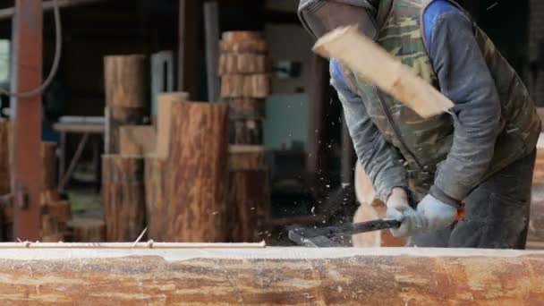 Trabajador de la construcción corta una ranura en el árbol para un futuro hogar. Máscara protectora y auriculares en la cabeza del constructor. Hangar con una parte de la futura casa en el fondo — Vídeos de Stock