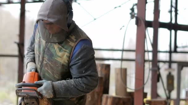 Bauarbeiter schneidet Holzkettensäge für künftiges Eigenheim Schutzmaske und Kopfhörer auf dem Kopf des Bauarbeiters und jede Menge Sägemehl. Hangar mit einem Teil der zukünftigen Heimat im Hintergrund — Stockvideo