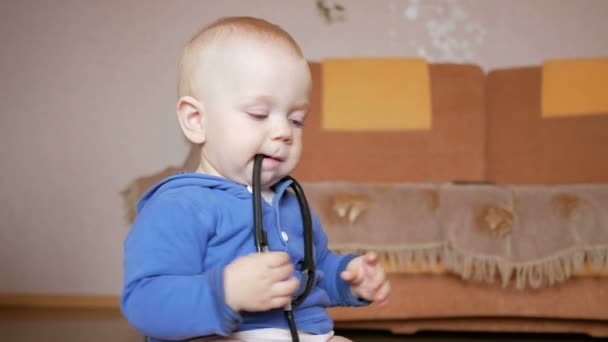 Милая малышка со стетоскопом дома. Тоддлер жует зубы медицинского устройства — стоковое видео
