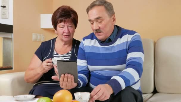 Boldog idősebb pár lelkesen nézett képernyő tabletta számítógép, egy csésze tea. Férj és feleség beszélünk, amit láttak — Stock videók
