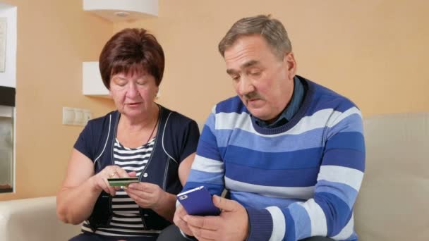 Couple de personnes âgées faisant leurs achats dans la boutique en ligne par carte de crédit et téléphone. Homme et femme assis à la maison carte de données sont administrés soigneusement sur le canapé — Video