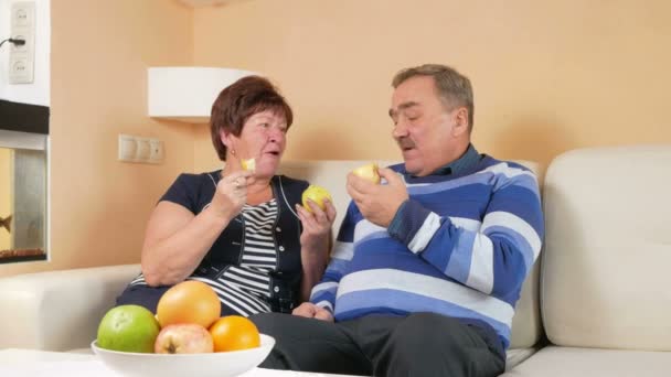 Starší pár, odpočívá doma na gauči a jíst jablka. Na stole je talíř z různých druhů ovoce. Muž a žena vzájemně komunikovat. — Stock video